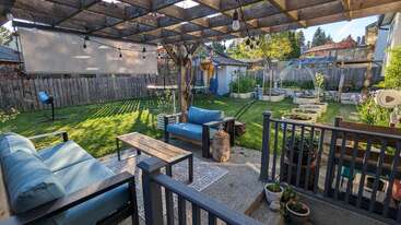Der gemütliche Garten hat eine Pergola mit Hängelampen, bequemen Sitzgelegenheiten, Topfpflanzen, einem gepflegten Rasen, Hochbeeten, einem Schuppen und einem Holzzaun.