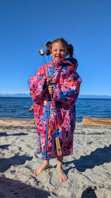 Ein lächelndes Kind steht barfuß an einem Sandstrand, trägt eine bunte, übergroße Jacke und hält geröstete Marshmallows in der Hand, während sich hinter ihm das Meer und der strahlend blaue Himmel erstrecken.
