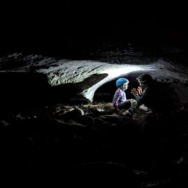 Zwei Leute, darunter ein Kind mit blauem Helm, sitzen in einer dunklen Höhle, beleuchtet von einer Taschenlampe, und quatschen oder genießen einfach den Moment in dieser geheimnisvollen Umgebung.