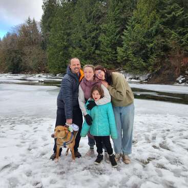 Eine glückliche vierköpfige Familie, eingemummt in Winterjacken, steht zusammen auf schneebedecktem Boden in der Nähe eines gefrorenen Flusses, zusammen mit ihrem braunen Hund, umgeben von grünen Bäumen.