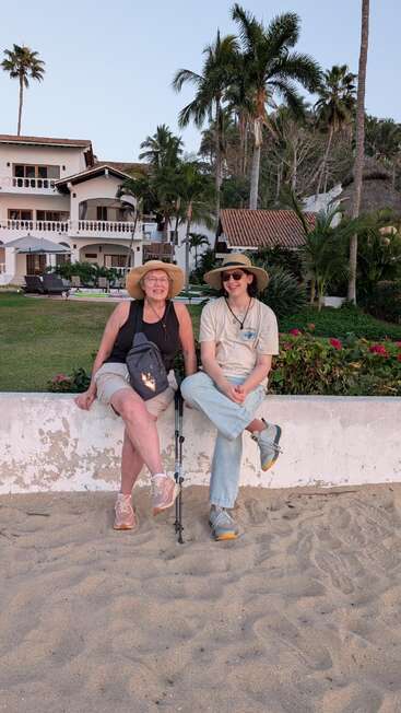 Two people wearing hats sit on a low wall by the beach, smiling. There are palm trees, a garden, and white buildings in the background.