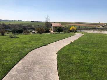 Un sendero de hormigón sinuoso serpentea a través de un césped verde y bien cuidado, y conduce a un pequeño edificio. Los campos del campo y el cielo azul y despejado completan esta tranquila estampa.