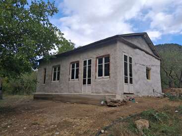 Ein kleines, rustikales Haus mit weißen Wänden und vielen Fenstern steht in einer ländlichen Umgebung, umgeben von Bäumen und Natur, unter einem teilweise bewölkten Himmel.