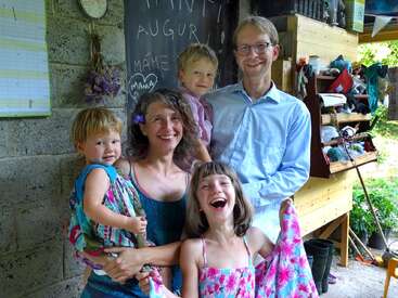 A happy family of five poses together, smiling warmly. The background features a chalkboard with writing and hearts, shelves, and hanging lavender. Casual, cheerful atmosphere.
