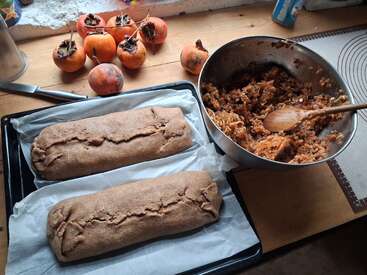 On a wooden countertop, two unbaked loaves rest on a baking tray. Beside them, a mixing bowl contains persimmon filling, with fresh persimmons nearby. Cozy kitchen scene.