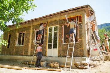 Um grupo de pessoas trabalha junto na construção ou reforma de uma casinha, aplicando materiais nas paredes externas usando escadas, cercadas por árvores e sob um sol quente.