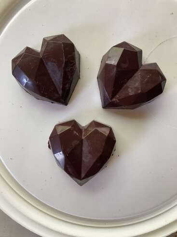 Tres corazones de chocolate negro con un diseño geométrico facetado descansan sobre un plato blanco, reflejando suavemente la luz. El conjunto es sencillo y evoca un aire moderno y elegante.
