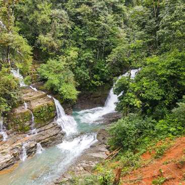A serene waterfall cascades over rocky ledges, surrounded by lush green foliage and dense forest, creating a tranquil, natural paradise in a vibrant jungle setting.