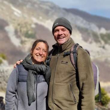 A imagem mostra um homem e uma mulher juntos em frente a um cenário montanhoso, com o braço do homem ao redor do ombro da mulher.