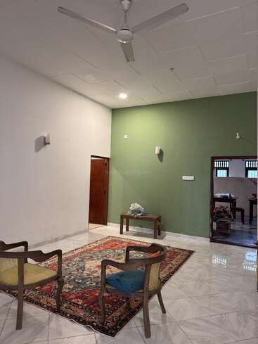 Esta imagem mostra uma sala de estar simples com duas cadeiras de madeira, um tapete colorido, uma pequena mesa, uma parede verde de destaque, um ventilador de teto branco e um piso de ladrilhos.