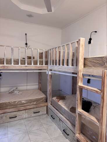 Esta imagem mostra um quarto pequeno e limpo com quatro beliches de madeira rústica dispostos em forma de L, gavetas de armazenamento embaixo e luzes de leitura individuais montadas na parede.