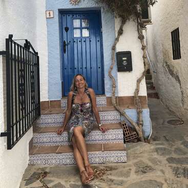 Une femme est assise, souriante, sur des marches décoratives en carrelage devant une charmante porte bleue, entourée de murs blancs et d'éléments rustiques, dégageant une atmosphère paisible et détendue.