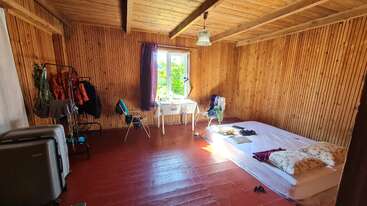 This cozy wooden room features a simple mattress on the floor, a small table by a sunlit window, clothes rack, red floor, and minimal furniture.