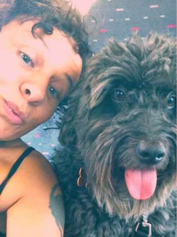 La imagen muestra a una mujer de pelo corto y rizado posando para un selfie con un perro negro de tamaño mediano con la lengua fuera.