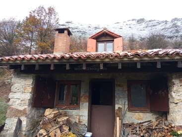 Una rústica cabaña de piedra con tejado de tejas rojas y chimenea, rodeada de árboles y leña, se asienta bajo una colina nevada en un entorno tranquilo.