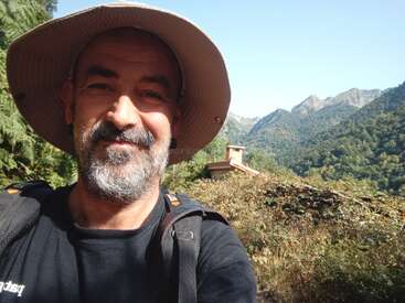 Un hombre con barba y sombrero sonríe para hacerse un selfie al aire libre. Está rodeado de exuberantes montañas verdes, follaje y un cielo azul claro, disfrutando de la naturaleza.