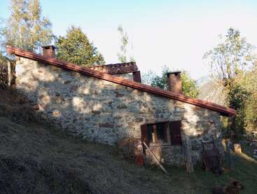 Una rústica casa de piedra con tejado inclinado de tejas rojas se asienta en una ladera cubierta de hierba, rodeada de altos árboles y montañas lejanas al fondo.