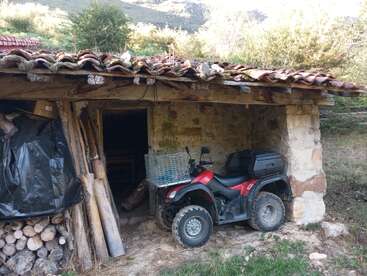 Un rústico cobertizo de piedra con tejado de tejas alberga leña apilada y un quad rojo aparcado con una cesta metálica, en medio de un paisaje rural y boscoso.