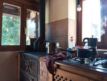 Esta acogedora cocina cuenta con una estufa de leña, ollas al fuego, una humeante cafetera moka, luminosas ventanas y una decoración rústica iluminada por la luz natural.