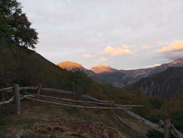 La foto muestra una rústica valla de madera en primer plano, con vistas a verdes colinas, lejanas montañas iluminadas por el sol, árboles y un colorido cielo con nubes dispersas al atardecer.