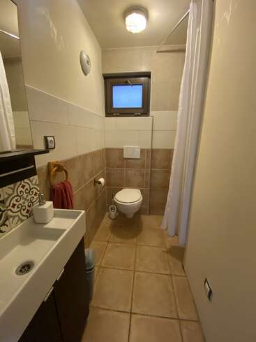 Cette photo montre une petite salle de bains avec du carrelage beige, des toilettes suspendues, un lavabo compact, un miroir, un porte-serviette avec une serviette rouge, une fenêtre, un rideau de douche et un plafonnier.