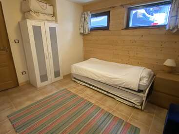 Cette chambre confortable comprend un lit simple, des murs en bois, un sol carrelé, un tapis rayé, une armoire surmontée de sacs de rangement et deux petites fenêtres ornées de rideaux.