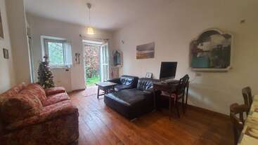 Un salon confortable avec du parquet, un canapé rouge à motifs, un canapé d'angle noir, un bureau avec un ordinateur, un sapin de Noël, des décorations murales et une porte donnant sur le jardin.