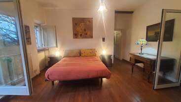 Une chambre confortable avec du parquet, un lit double avec des draps roses, des tables de chevet, un éclairage chaleureux, des œuvres d'art, des fenêtres ouvertes, un bureau, une lampe en vitrail et une atmosphère spacieuse.