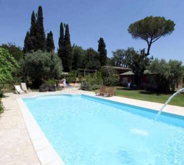Une piscine extérieure paisible entourée de chaises longues, d'arbres verdoyants et de buissons. Le ciel est clair et bleu, évoquant une atmosphère estivale paisible.