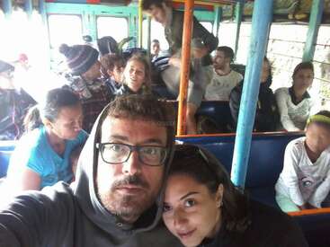 Un groupe de personnes est assis côte à côte dans un bus aux couleurs vives. Deux personnes au premier plan prennent un selfie, les autres semblent détendues et discutent tranquillement.
