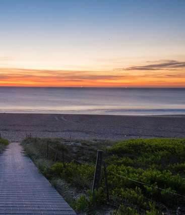 Das Bild zeigt eine ruhige Strandszene bei Sonnenuntergang mit einem gepflasterten Weg, üppigem Grün und einem weiten Meer mit einem leuchtenden Himmel und sanften Wellen.