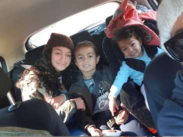 Das Bild zeigt drei Kinder, die auf dem Rücksitz eines Autos sitzen, mit einer Frau auf der linken Seite und zwei Kindern in Autositzen auf der rechten Seite.