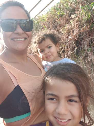 Das Bild zeigt eine Frau und zwei junge Mädchen, die für ein Selfie vor einer Wiese mit einem Zaun posieren. Die Frau trägt eine Sonnenbrille.
