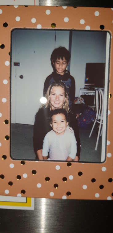 Das Bild zeigt ein Foto von einer Frau und zwei Kindern, die für ein Foto posieren, wobei die Frau links und die Kinder rechts stehen. Der Hintergrund ist unscharf.