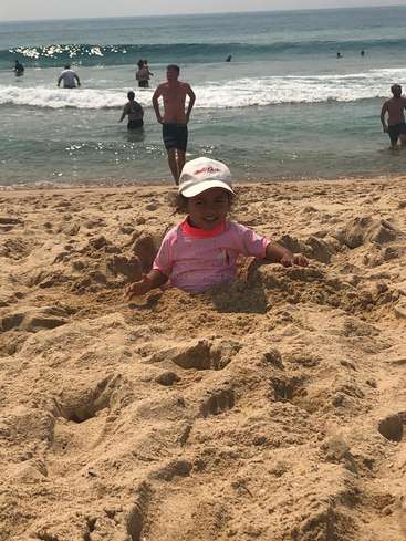 Ein junges Mädchen mit rosa Hemd und weißer Mütze liegt an einem Strand im Sand, während hinter ihr Menschen im Meer schwimmen.