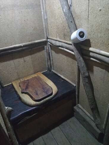 This image shows a rustic, wooden toilet inside a small, simple structure. The toilet seat is made of wood, with a roll of toilet paper on a branch.