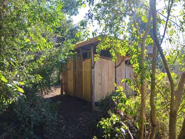 A small wooden shed is nestled among lush, leafy green trees. Sunlight filters through the foliage, creating a peaceful, natural atmosphere around the rustic structure.
