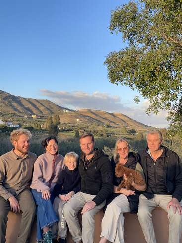 Six personnes et un chien sont assis en plein air sur un muret et sourient. Derrière eux se trouvent des collines verdoyantes, des maisons éparpillées et un ciel bleu clair.