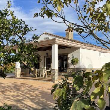L'image représente une maison à un étage avec un patio et un arbre au premier plan, sur fond de ciel bleu avec des nuages.