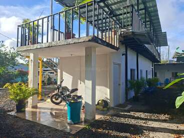 Un bâtiment blanc de deux étages, dont le balcon supérieur est orné de plantes en pot, avec un patio couvert en contrebas, une moto garée et une végétation tropicale tout autour.