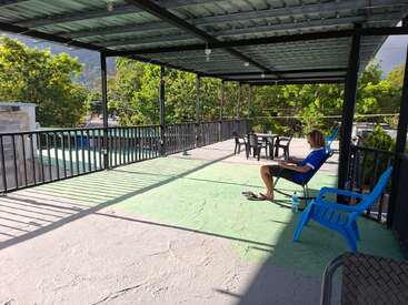 Une personne est assise sur une chaise bleue sous un auvent, devant son ordinateur portable. La lumière du soleil projette des ombres. On aperçoit des arbres et des montagnes à l'arrière-plan. L'atmosphère est paisible.