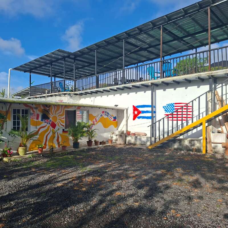 Une cour ensoleillée agrémentée de plantes en pot, des fresques murales colorées représentant des drapeaux, notamment ceux de Cuba et des États-Unis, ainsi qu'un bâtiment doté d'une terrasse à l'étage, sous un toit métallique.