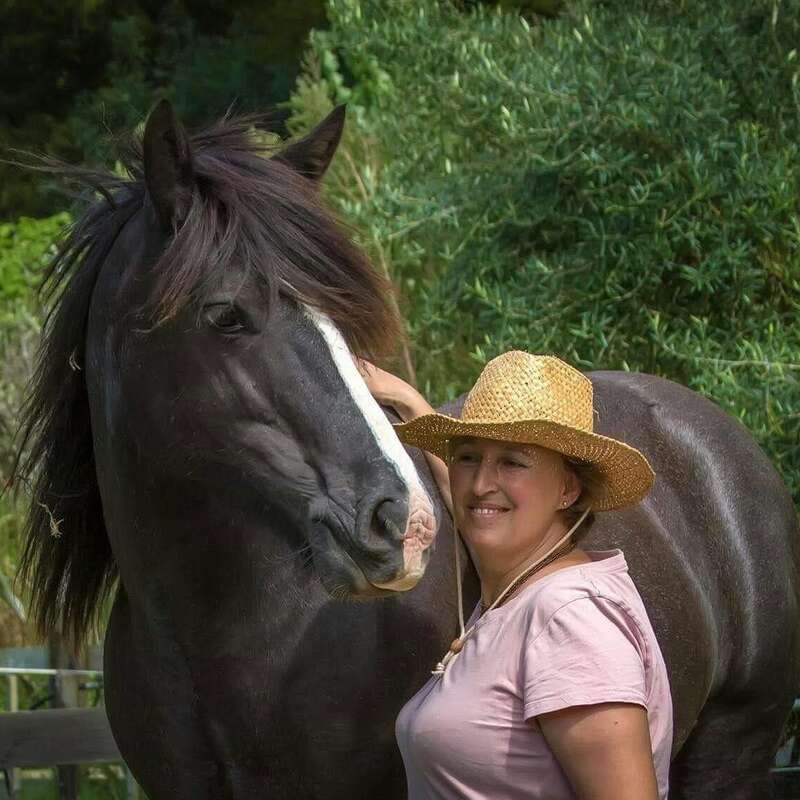 Uma mulher sorridente com um chapéu de palha está ao lado de um grande cavalo escuro com uma faixa branca. Uma folhagem verde exuberante os rodeia, criando um ambiente natural e tranquilo.