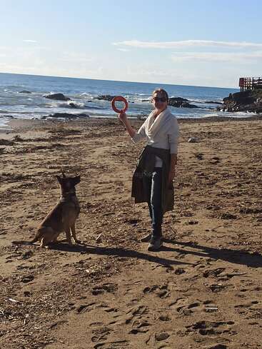 Uma mulher está em pé numa praia arenosa segurando um brinquedo vermelho em forma de anel, enquanto um cão senta-se atentamente à sua frente, com o mar atrás deles.