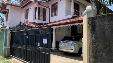 Uma casa de dois andares com telhado de telhas vermelhas, paredes brancas e varandas. Portão preto ornamentado na frente, entrada da garagem com um carro prateado estacionado dentro, dia ensolarado.