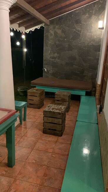 Um pátio coberto aconchegante tem bancos e mesas azul-petróleo, banquetas rústicas de madeira, piso de ladrilho, uma espreguiçadeira almofadada, luzes de corda penduradas e uma parede de pedra.