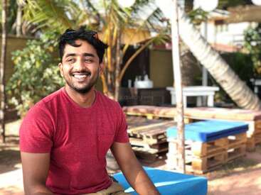 A smiling man in a red t-shirt sits outdoors on a bright sunny day. Palm trees and wooden pallet benches create a relaxed, tropical vacation atmosphere behind him.