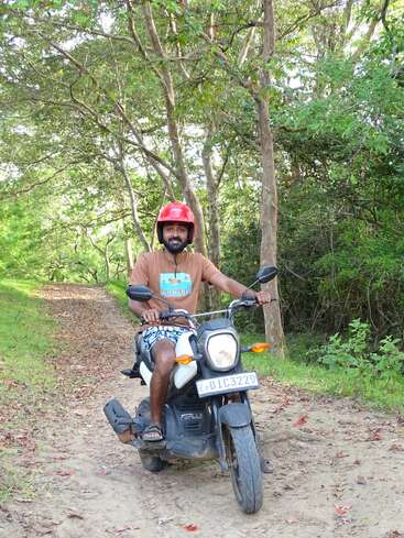 Um homem usando capacete vermelho e camisa marrom dirige uma scooter em um caminho de terra cercado por árvores e arbustos verdes, desfrutando de um ambiente externo tranquilo.