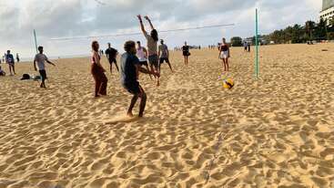 Um grupo de pessoas está jogando vôlei de praia em um dia ensolarado. Alguns pulam para acertar a bola. A atmosfera parece animada e divertida, com muitos participantes.