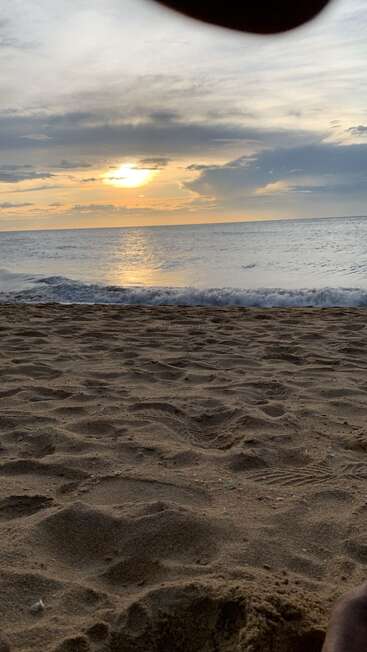 Uma praia de areia se estende em direção a ondas calmas enquanto o sol se põe atrás de nuvens esparsas, lançando um reflexo dourado e quente na água tranquila do oceano. Noite tranquila.